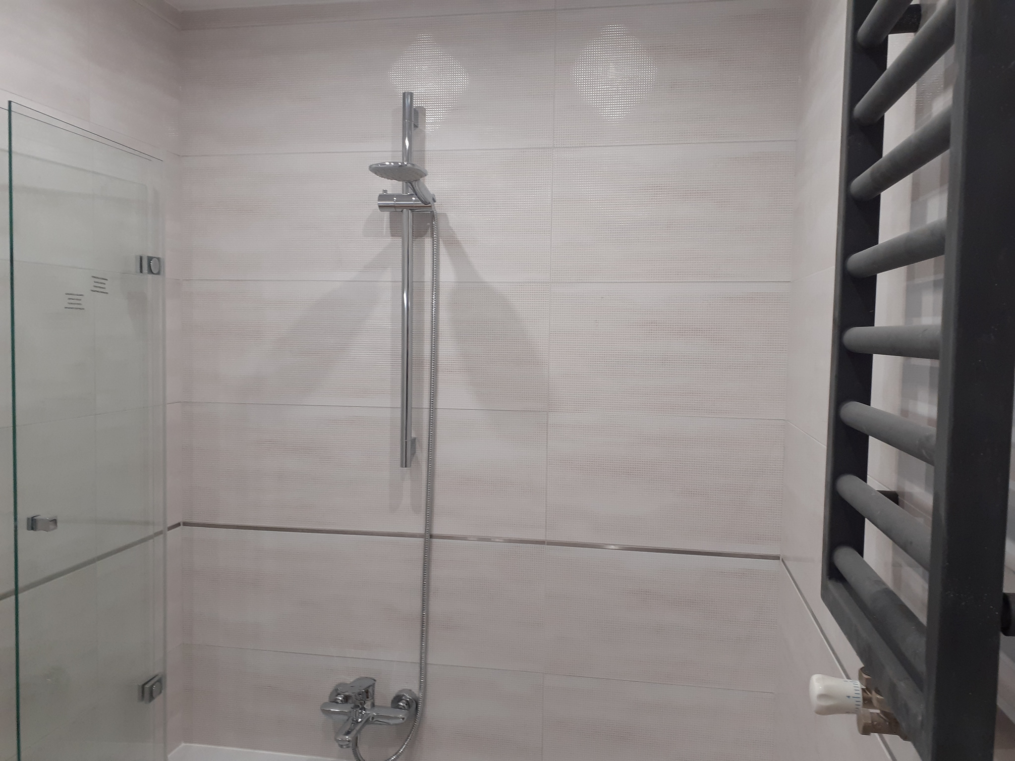 Wykończona łazienka z kabiną prysznicową ze szklanymi drzwiami, chromowaną armaturą, grzejnikiem drabinkowym w kolorze antracytowym i beżowymi płytkami z delikatną fakturą.