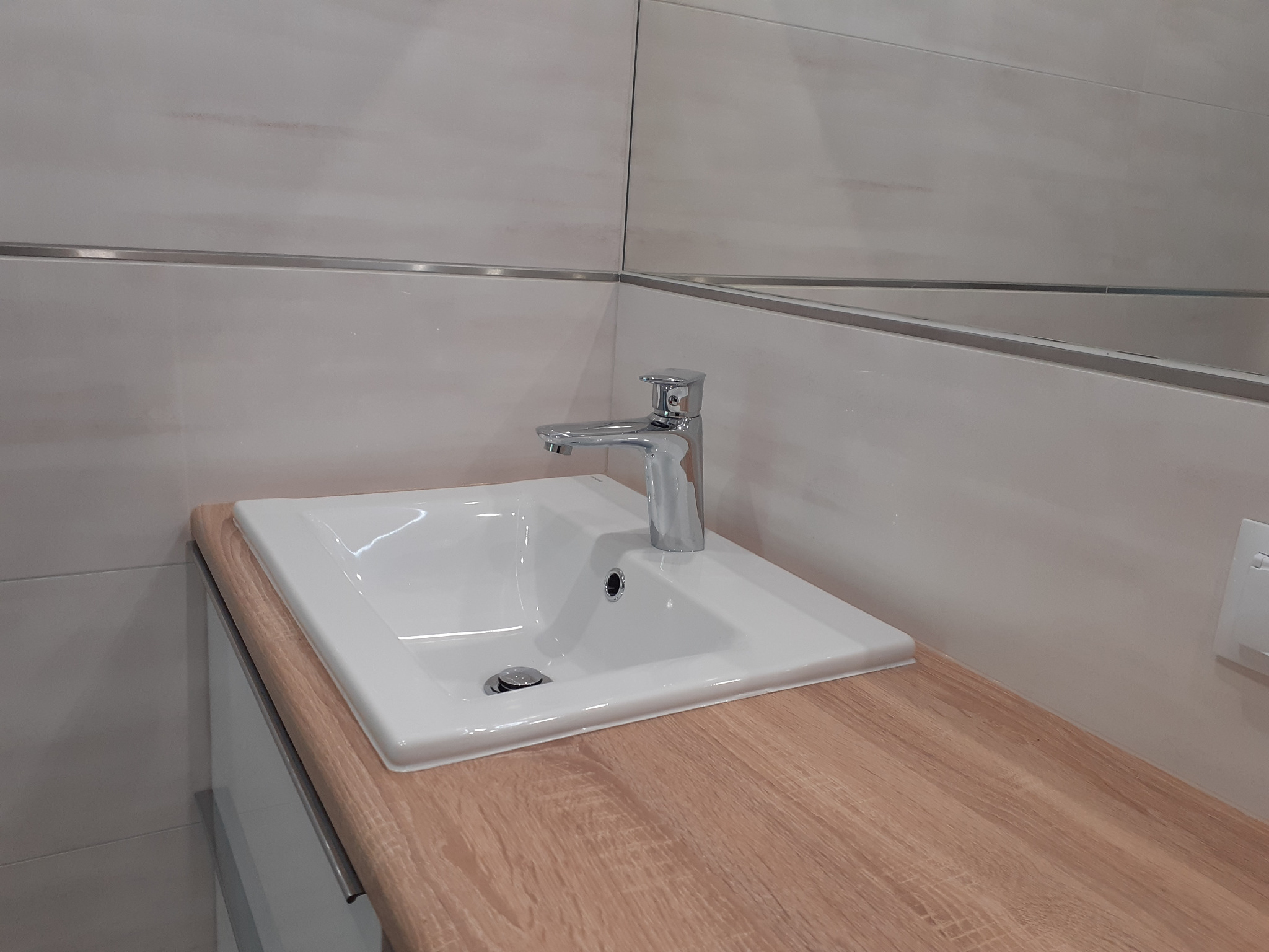 Biała umywalka z chromowaną baterią na blacie imitującym jasne drewno, z jasnymi płytkami na ścianie i lustrem w tle.