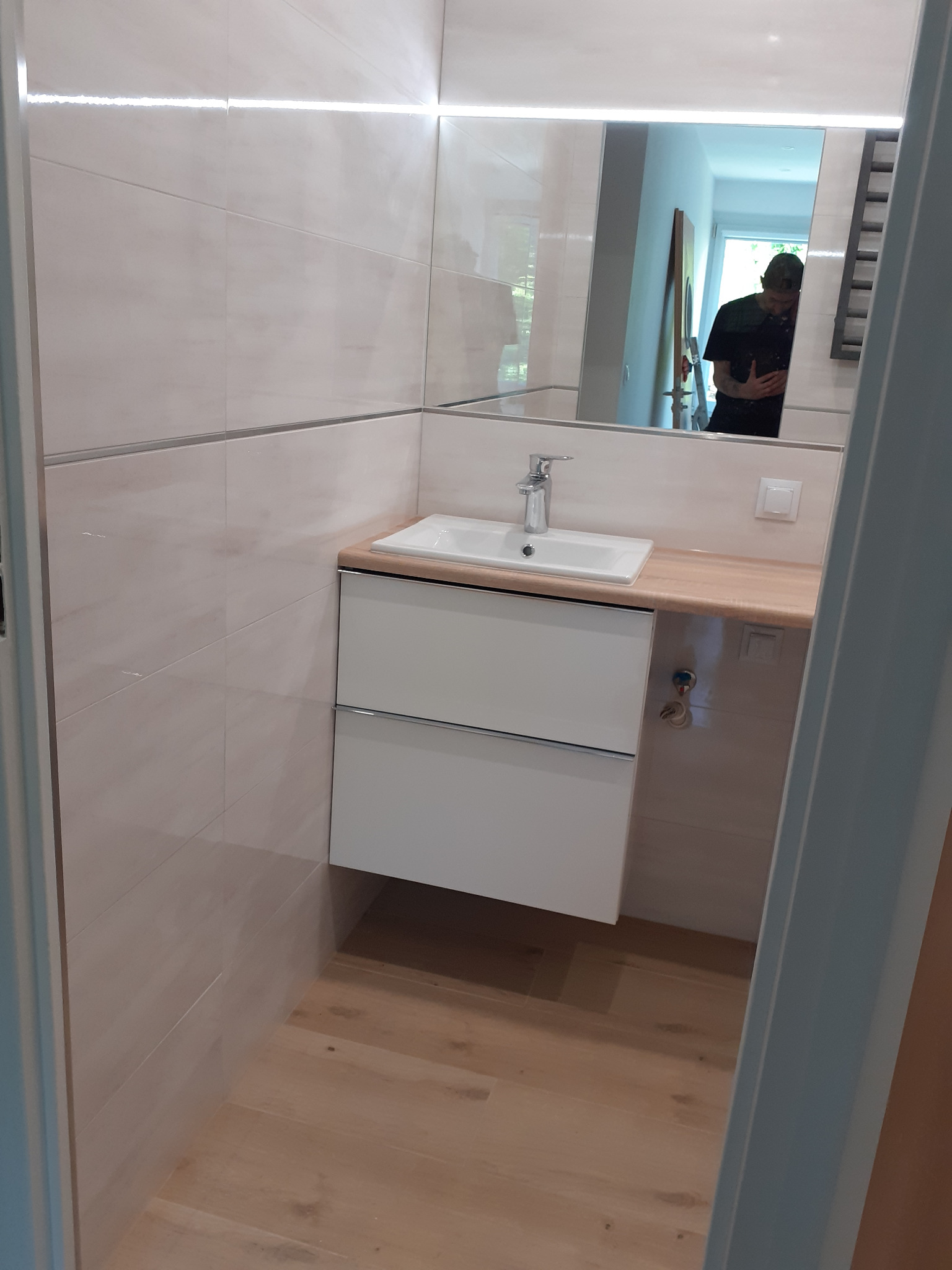 Nowoczesna łazienka z beżowymi płytkami, białym umywalką z szafką podwieszaną i drewnianym blatem, oświetleniem LED nad lustrem oraz drewnianą podłogą. W lustrze odbija się osoba.