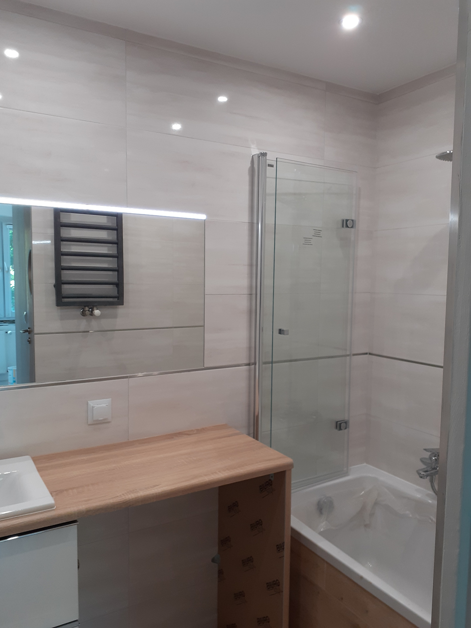 Jasna łazienka po remoncie z kabiną prysznicową ze składanymi drzwiami, umywalką z drewnianym blatem i grzejnikiem drabinkowym widocznym w lustrze.