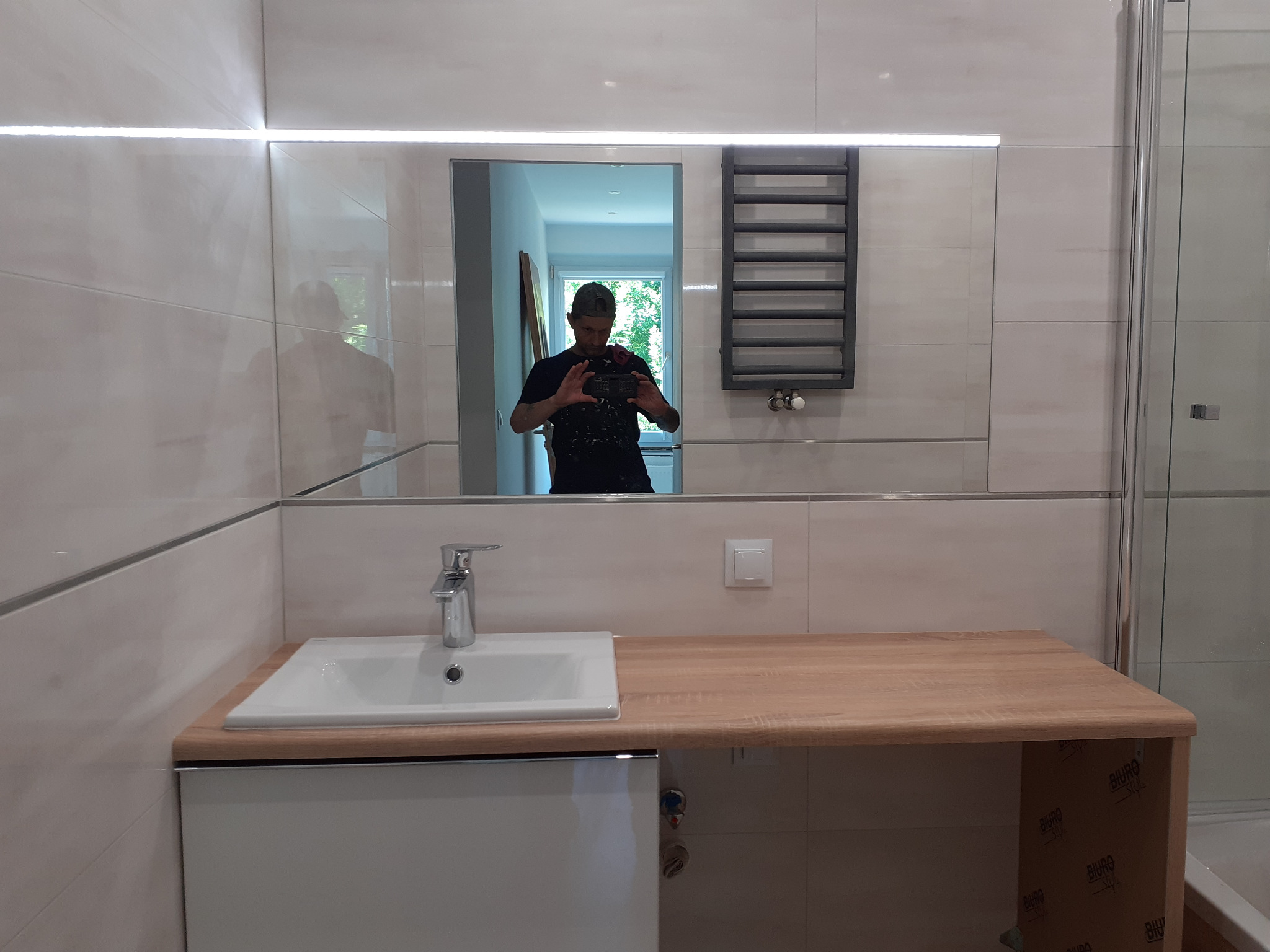 Nowoczesna łazienka z umywalką nablatową, drewnianym blatem, grzejnikiem drabinkowym i lustrem z podświetleniem LED, widoczny refleks fotografa w lustrze.