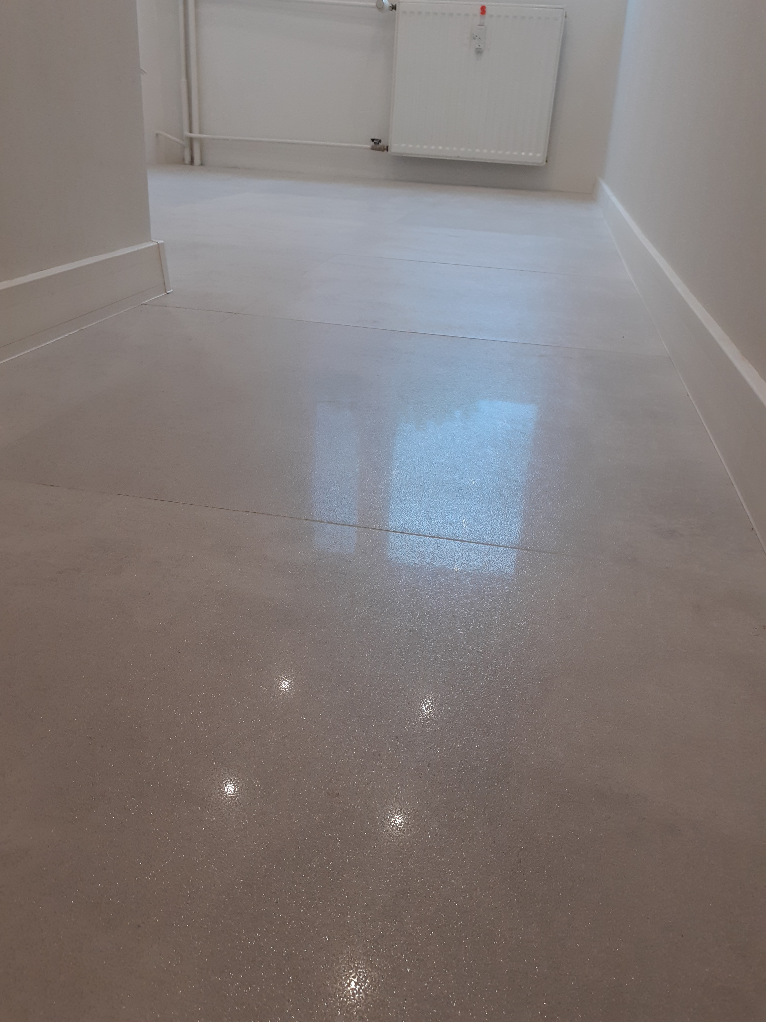 Szlifowana, beżowa posadzka z widocznymi szczelinami dylatacyjnymi i odblaskami światła, listwy przypodłogowe oraz biały grzejnik w tle.