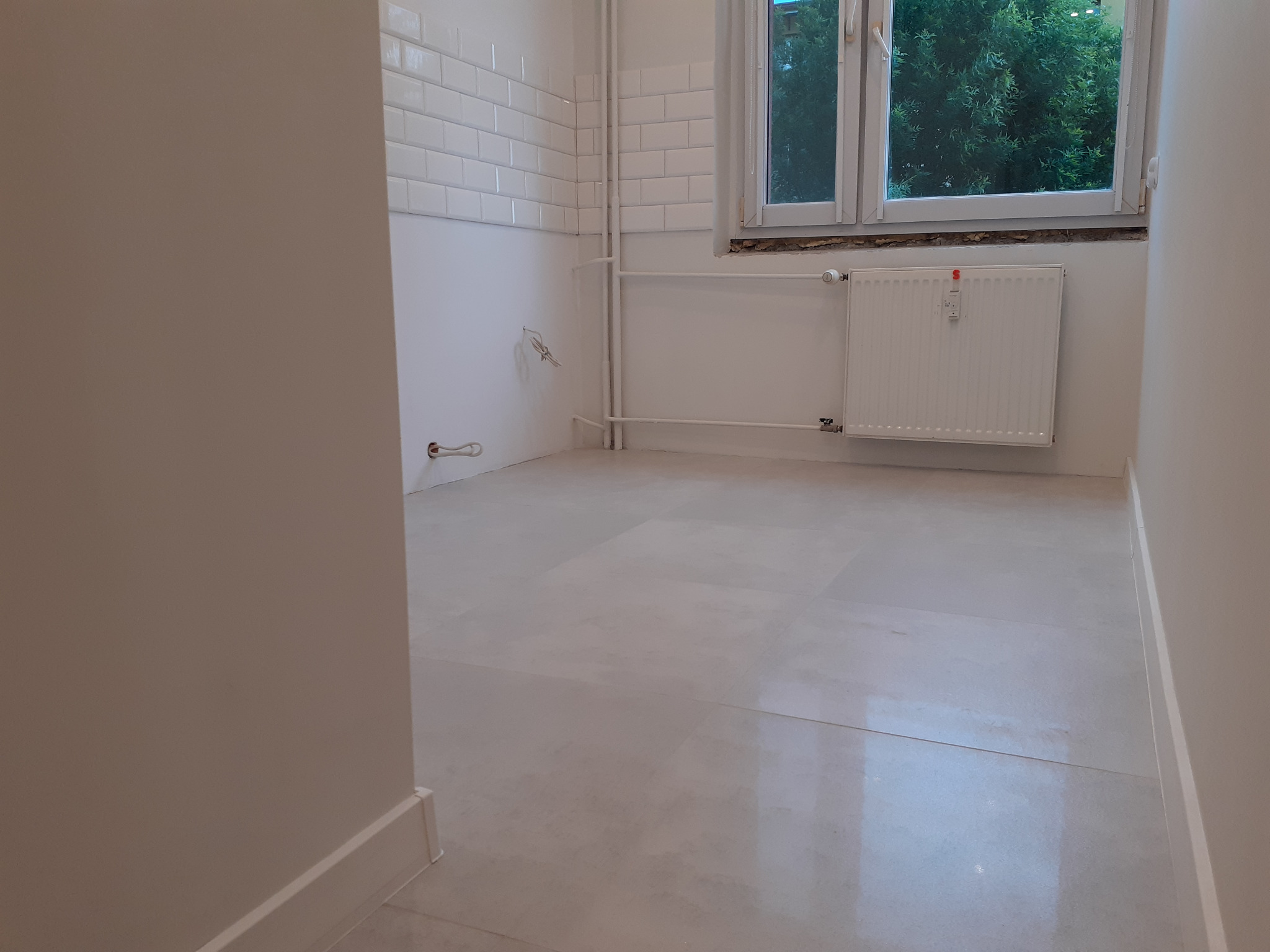 Wnętrze łazienki w trakcie remontu, widoczne białe płytki na ścianach, grzejnik, okno z widokiem na zieleń oraz jasna, świeżo wylana posadzka.