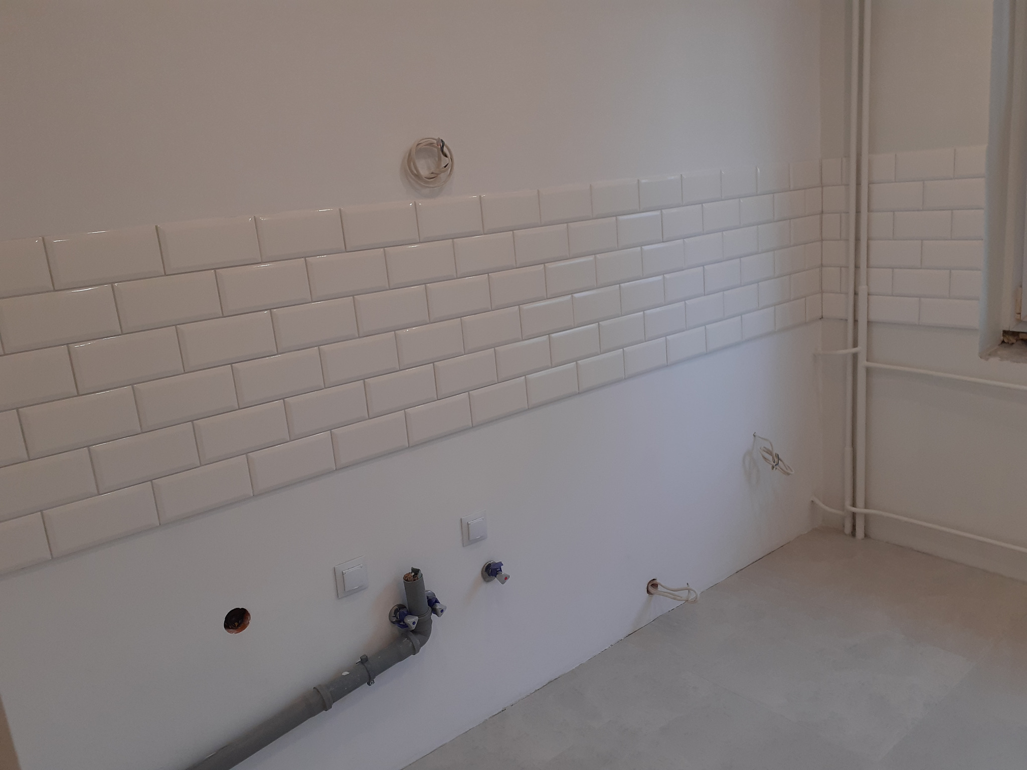 Świeżo położone, białe, prostokątne płytki na ścianie w kuchni w Rybniku, widoczne przyłącza wodne i elektryczne przed montażem armatury.