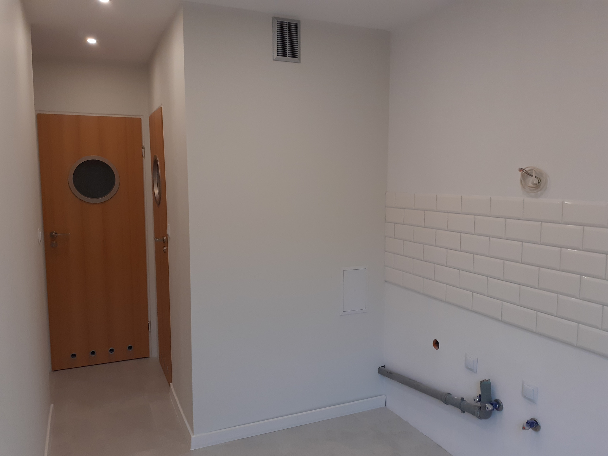 Wnętrze łazienki w trakcie prac wykończeniowych: białe, prostokątne płytki częściowo ułożone, widoczne rury instalacji wodnej i odpływowej, drzwi z okrągłym okienkiem w tle.