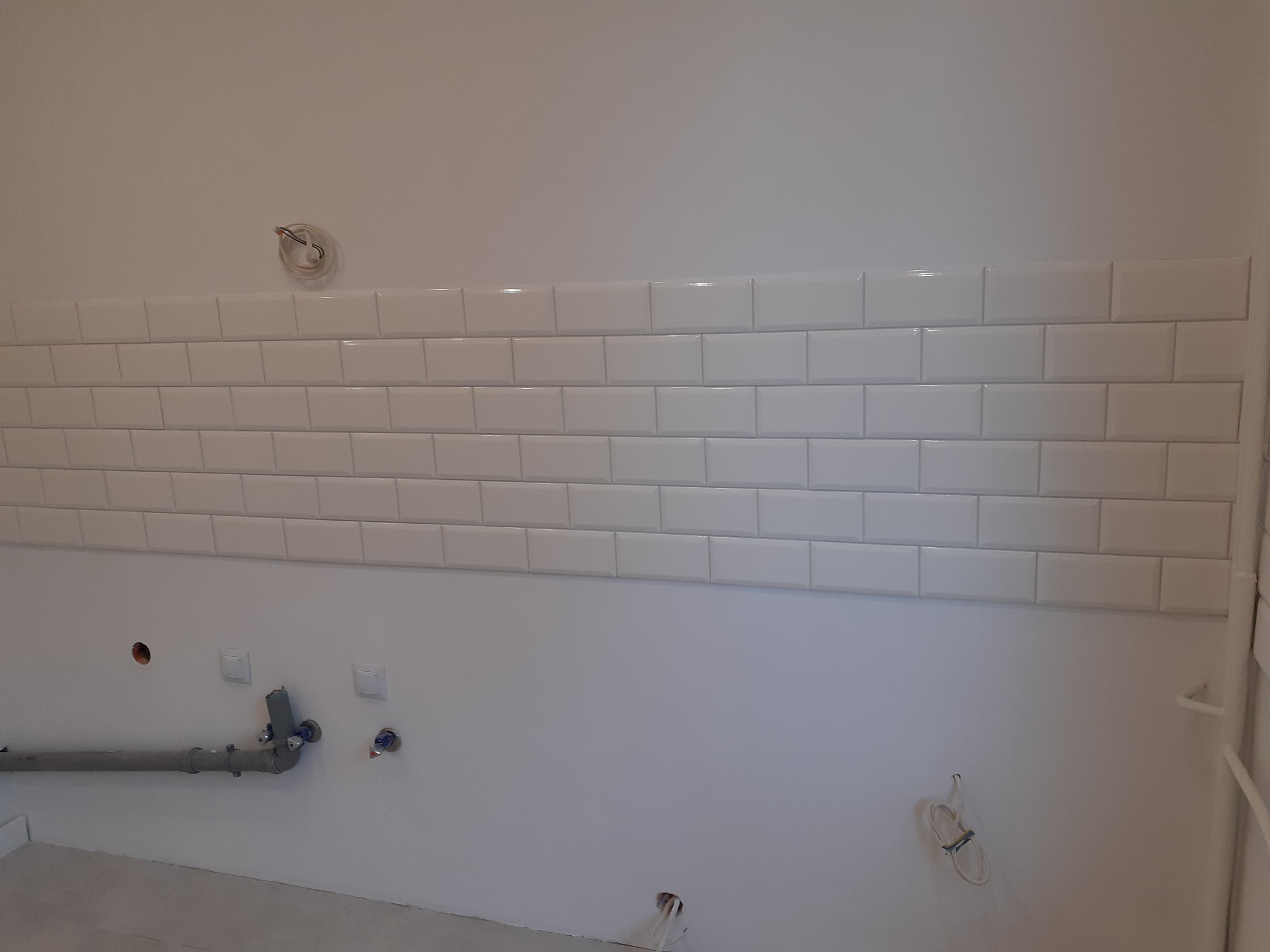 Ściana łazienki w trakcie remontu z ułożonymi białymi, podłużnymi płytkami w stylu metro, widoczne instalacje hydrauliczne i elektryczne przed ostatecznym wykończeniem.