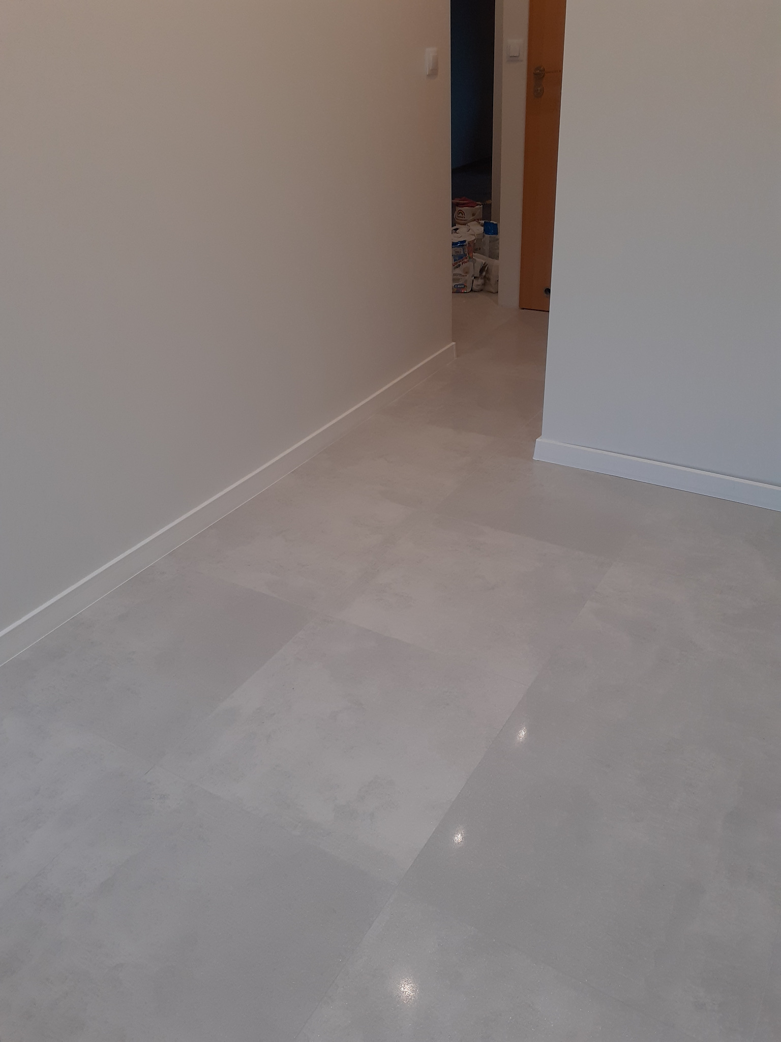 Jasnoszara, matowa podłoga z płytek ceramicznych w nowo wyremontowanym pokoju, widoczne listwy przypodłogowe i drzwi do kolejnego pomieszczenia, w którym widać puszki z farbą.