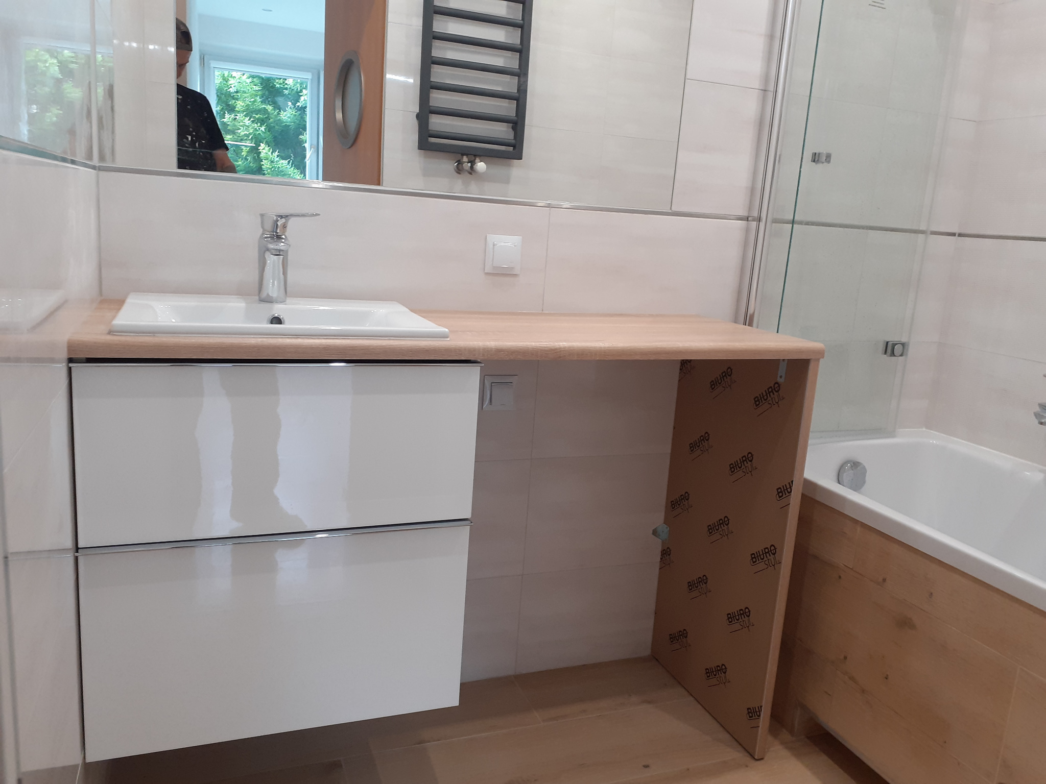 Nowoczesna łazienka z białymi płytkami, drewnianym blatem pod umywalką i kabiną prysznicową ze szklanymi drzwiami. Widoczne białe szafki pod umywalką, grzejnik drabinkowy na ścianie i okrągłe okno...