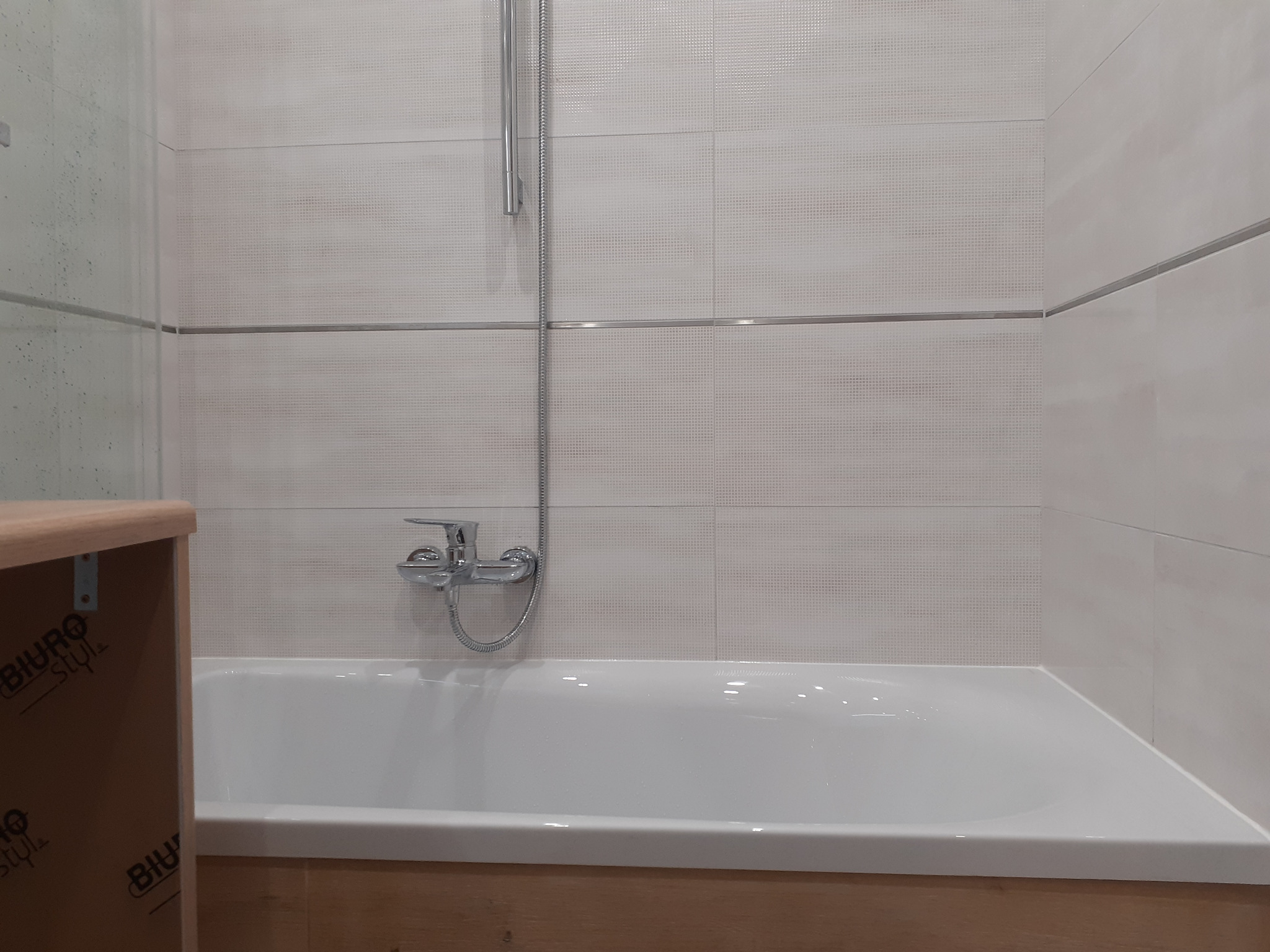 Biała wanna obudowana drewnopodobną płytą, chromowana bateria z prysznicem na tle jasnych, kwadratowych płytek z delikatnym wzorem.