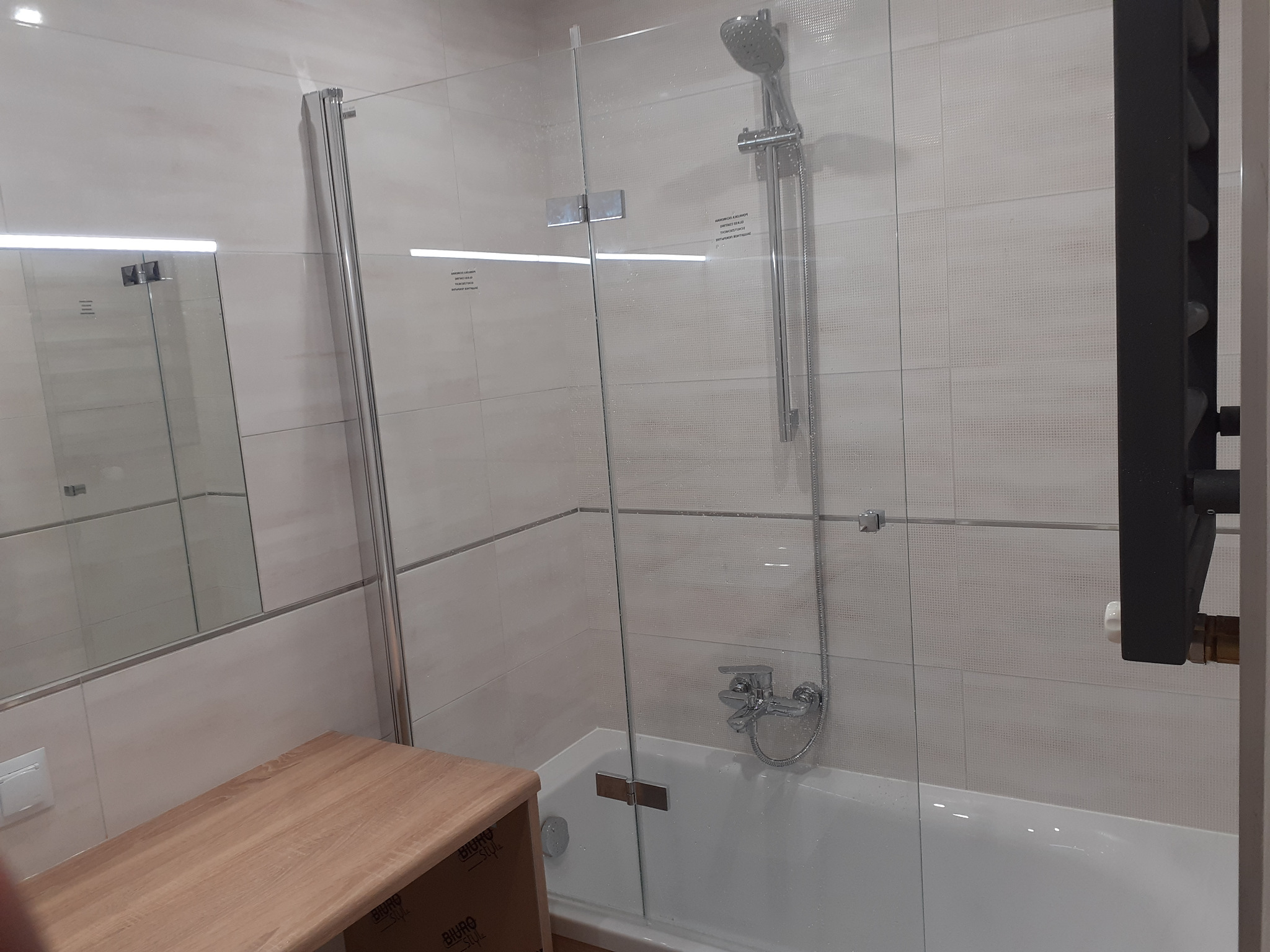 Nowoczesna kabina prysznicowa z elementami szklanymi i drewnianą szafką umywalkową, widok na baterię i grzejnik drabinkowy w łazience wykończonej jasnymi płytkami.