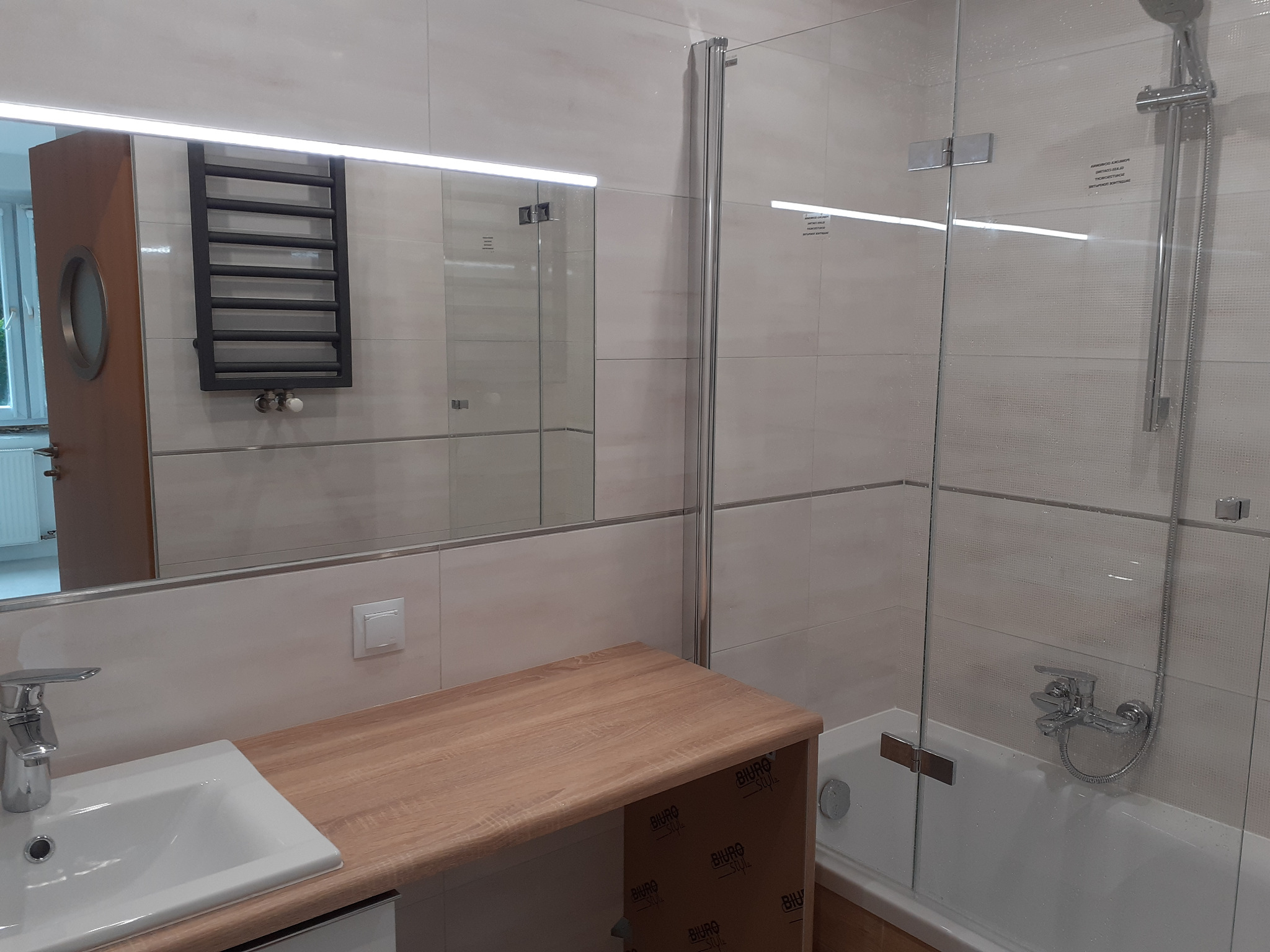 Nowoczesna łazienka z kabiną prysznicową z przezroczystego szkła, umywalką wpuszczaną w blat z drewnopodobnym wykończeniem, grzejnikiem drabinkowym i okrągłym oknem w drzwiach widocznym w lustrze.