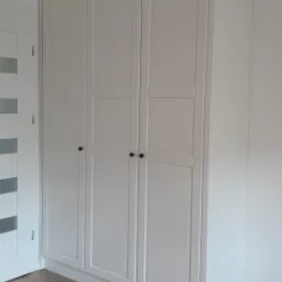 Biała, trzydrzwiowa szafa wnękowa z czarnymi gałkami, dopasowana do sufitu, na podłodze panele imitujące drewno, widoczny fragment białej ściany i drzwi z przeszkleniami.