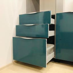 Dece - Meble - Niebiesko-szara szafka z trzema szufladami, z których dwie są lekko wysunięte, ukazując białe wnętrze, ustawiona w rogu pomieszczenia z beżowymi ścianami i podłogą.