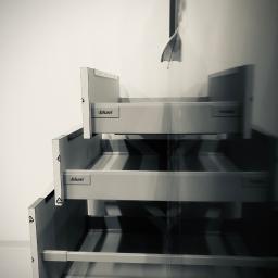 Dece - Meble - Częściowo zamontowane, szare szuflady z logo 'blum' ustawione jedna nad drugą, z widocznymi elementami mocującymi i symbolem recyklingu na boku, na tle białej ściany.