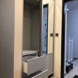 Dece - Meble - Wnętrze szafy wnękowej z otwartymi szufladami w jasnych odcieniach szarości i bieli, widoczne półki i drążek na ubrania, w tle drzwi do innego pomieszczenia i fragment schodów.