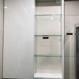 Biała szafka łazienkowa na wymiar z połyskującymi frontami i trzema szklanymi półkami, zamontowana na ścianie z płytek imitujących drewno.