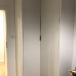 Biała szafa narożna z ciemnymi uchwytami, wkomponowana w narożnik pokoju, widoczna z bliska, z fragmentem podłogi i ściany z włącznikiem światła.