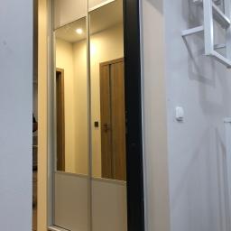 Dece - Meble - Wnętrze przedpokoju z dużą, przesuwną szafą z lustrzanymi drzwiami, odbijającymi fragment innego pokoju z drewnianymi drzwiami i dekoracją ścienną w tle.