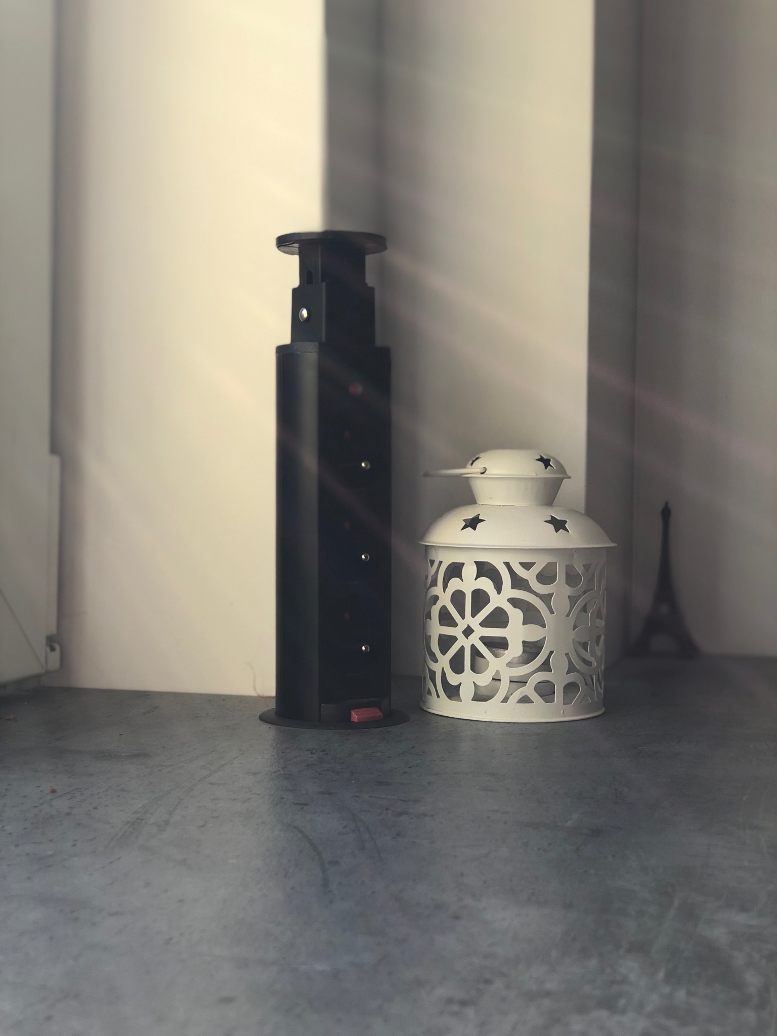 Czarne, metalowe podparcie blatu z regulacją wysokości stoi obok białego lampionu z wyciętymi wzorami na szarym blacie, z miniaturą wieży Eiffla w tle i promieniami słonecznymi.