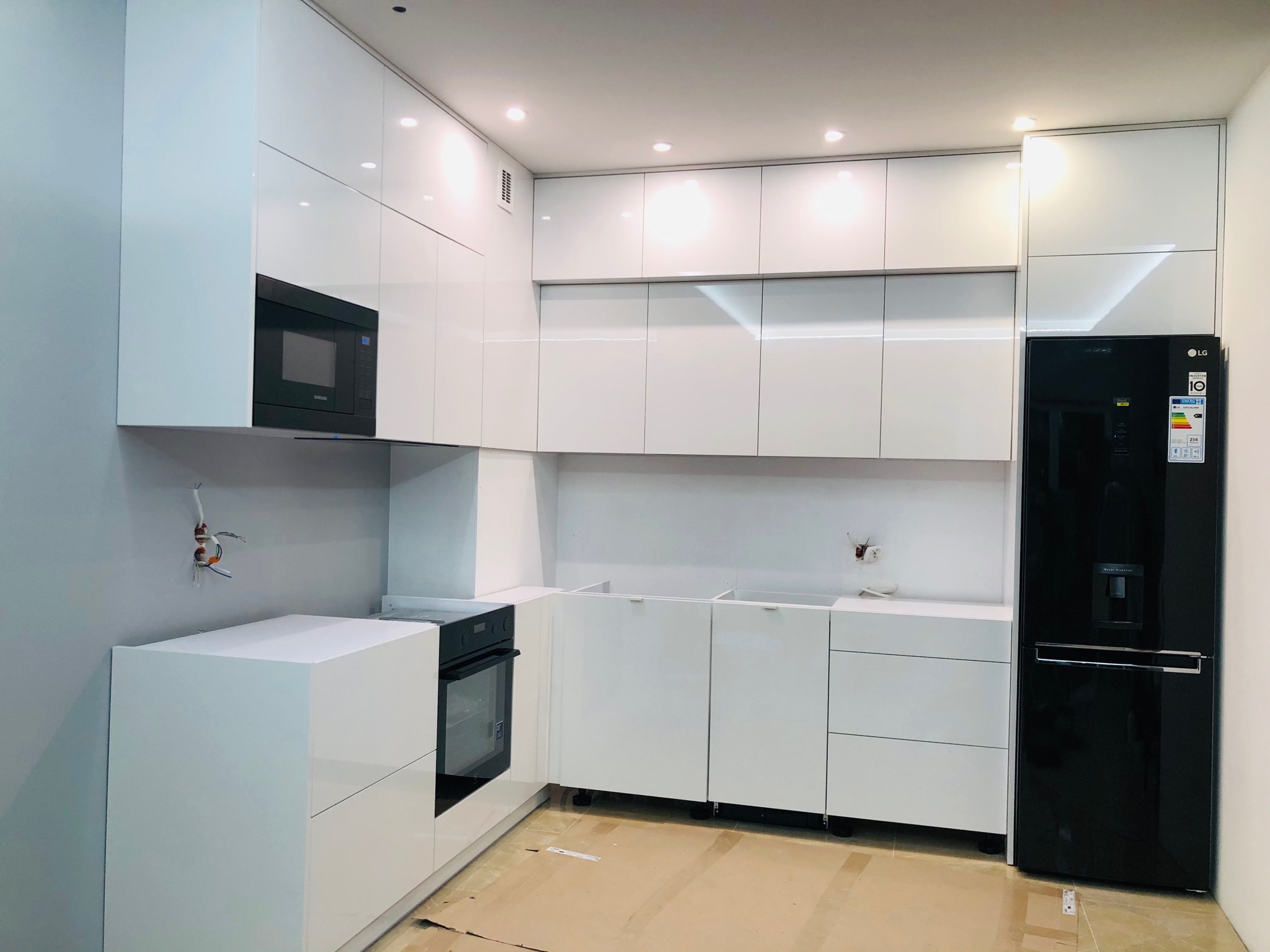 Nowoczesna, biała kuchnia z połyskiem w trakcie montażu, widoczne niedokończone podłączenia elektryczne, czarna lodówka LG w zabudowie.