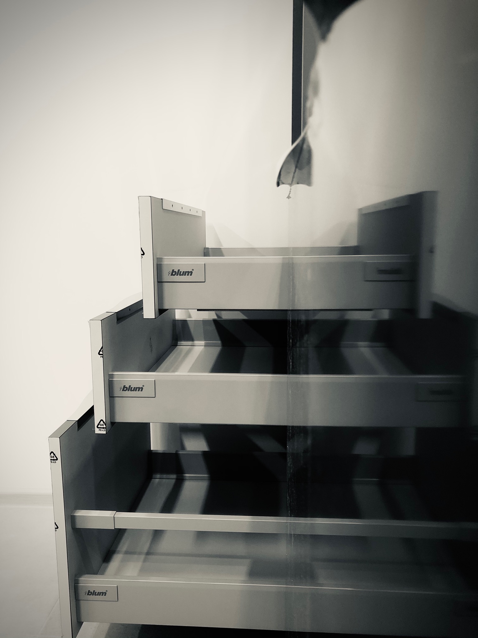 Częściowo zamontowane, szare szuflady z logo 'blum' ustawione jedna nad drugą, z widocznymi elementami mocującymi i symbolem recyklingu na boku, na tle białej ściany.