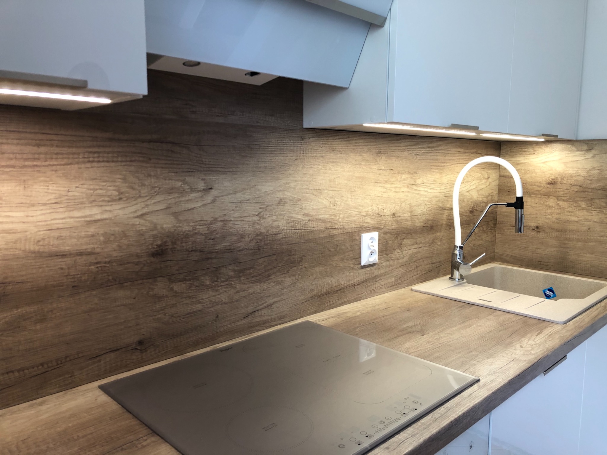 Nowoczesna kuchnia z drewnianym blatem, szarym zlewem i płytą indukcyjną; oświetlenie LED pod szafkami górnymi.