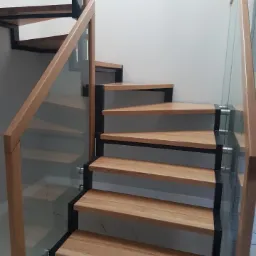 Nowoczesne schody z drewnianymi stopniami, czarną metalową konstrukcją i szklanymi balustradami, widoczne mocowania szkła do stopni.