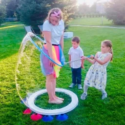 Dziewczyna w tęczowej spódnicy robi ogromne bańki mydlane na trawie, dwoje dzieci pomaga trzymając kijki, w tle zielony trawnik i drzewa.