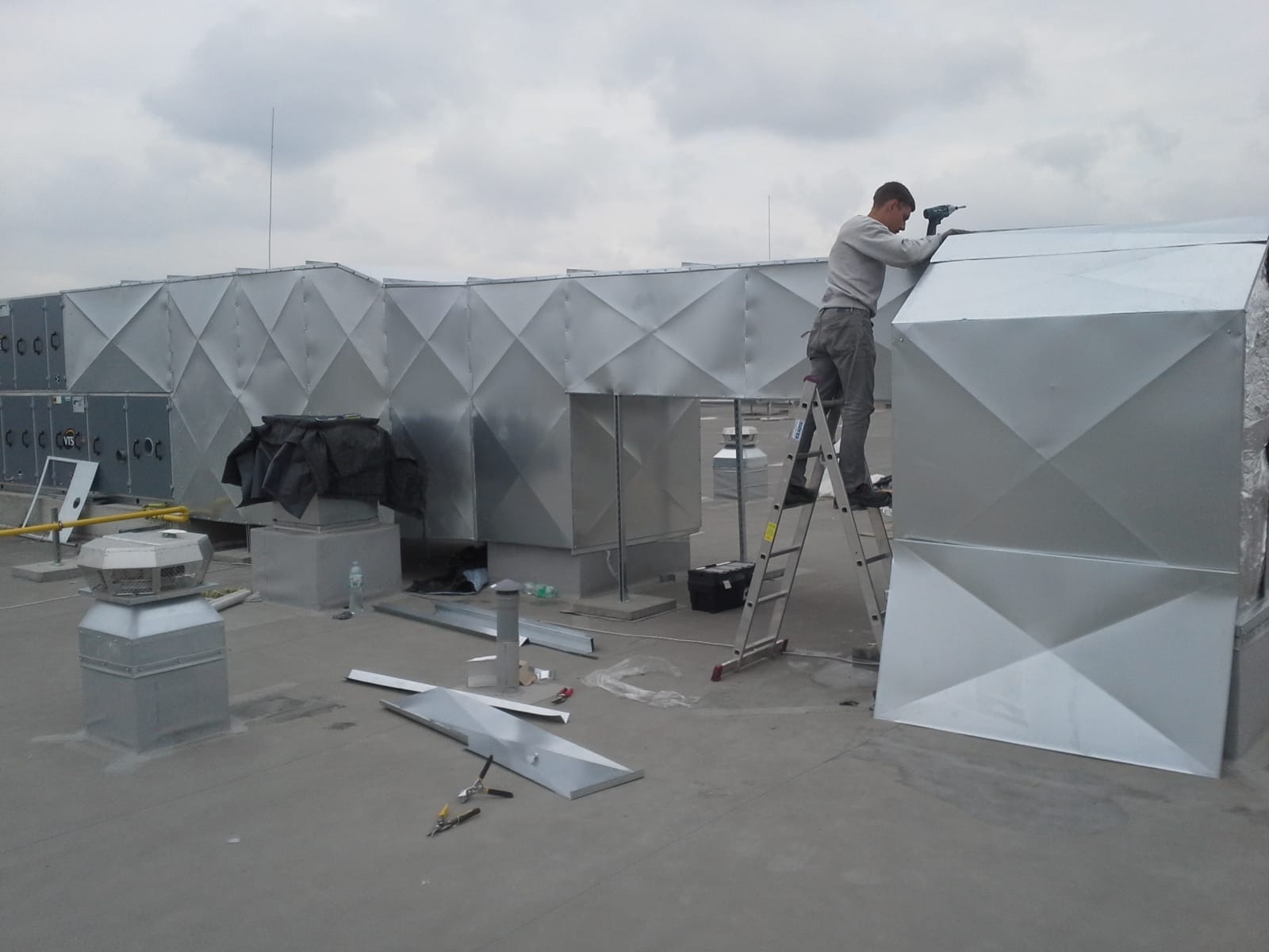 Pracownik na drabinie montuje srebrną instalację wentylacyjną na dachu budynku, widoczne narzędzia i elementy konstrukcyjne.
