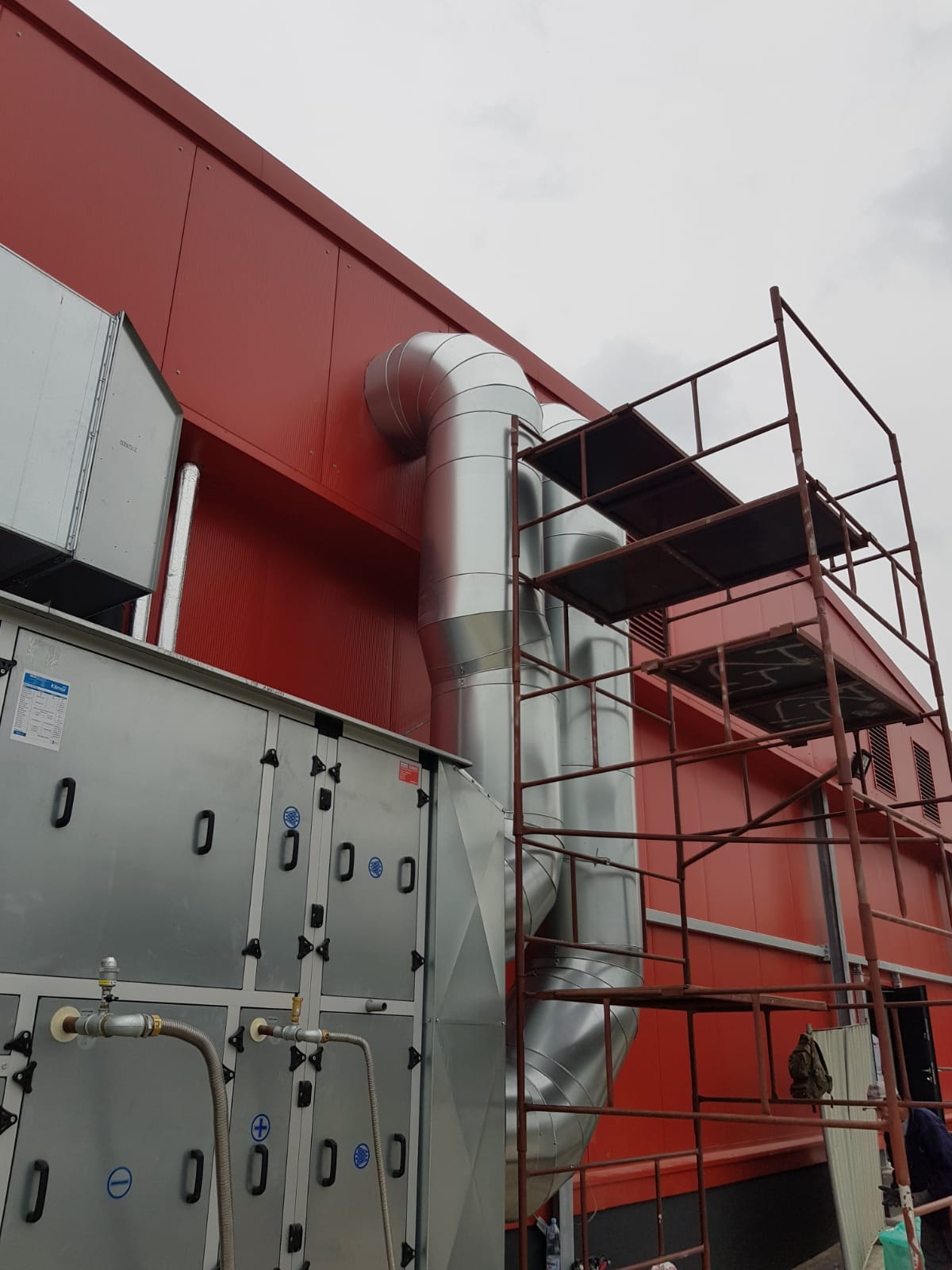 Zewnętrzny montaż systemu wentylacyjnego z rurami spiro na tle czerwonego budynku przemysłowego, widoczne rusztowanie i jednostka centralna z zaworami.