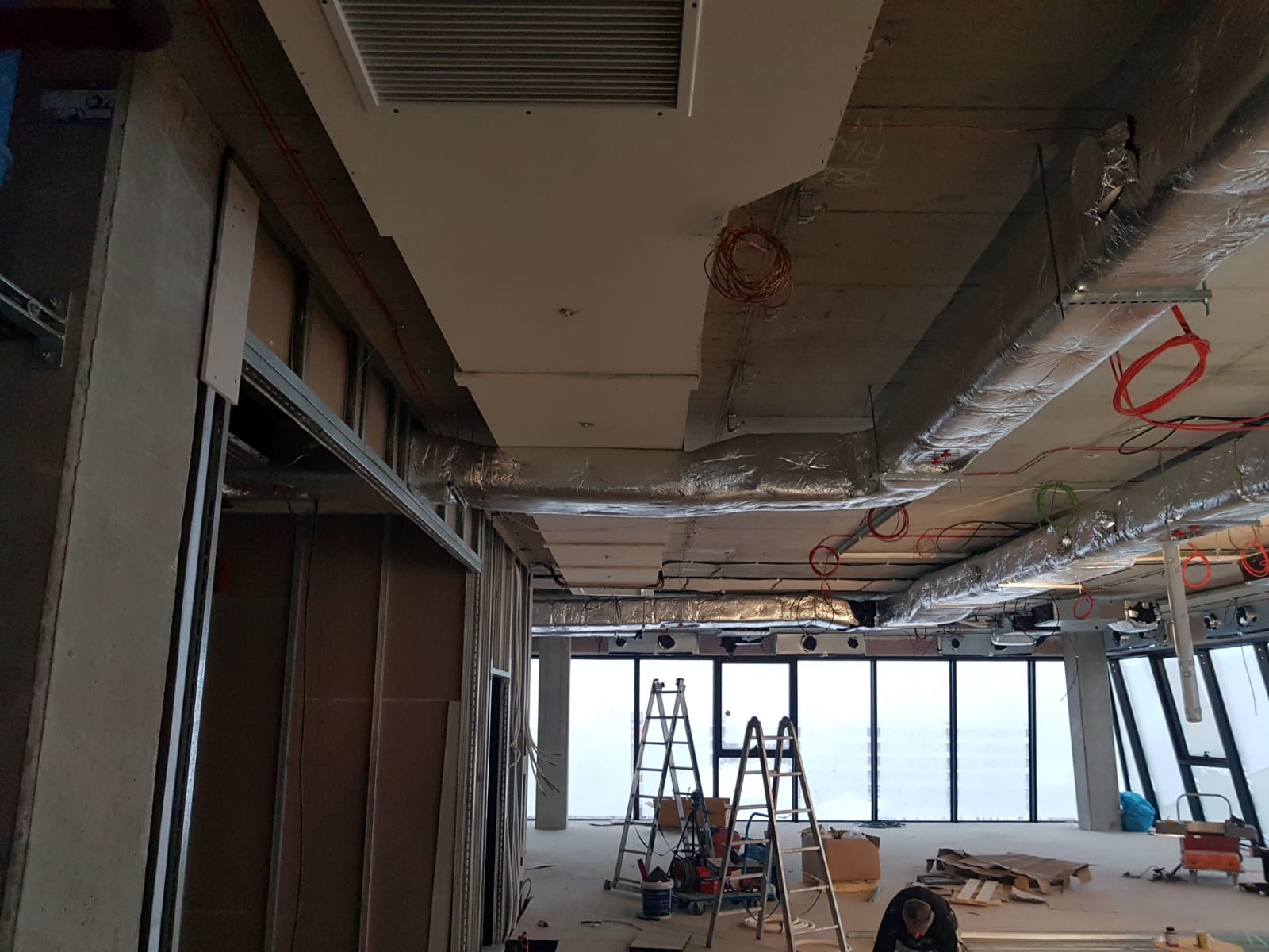 Wnętrze w trakcie montażu instalacji wentylacyjnej, widoczne kanały wentylacyjne pod sufitem, okablowanie, drabiny i elementy konstrukcyjne ścian.