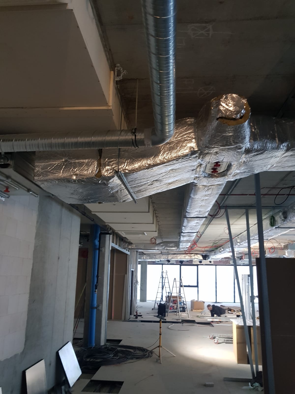 Montaż instalacji wentylacyjnej: widok na sufit z systemem rur wentylacyjnych w srebrnej izolacji, widoczny pracownik na drabinie w tle, surowy stan wykończenia wnętrza.