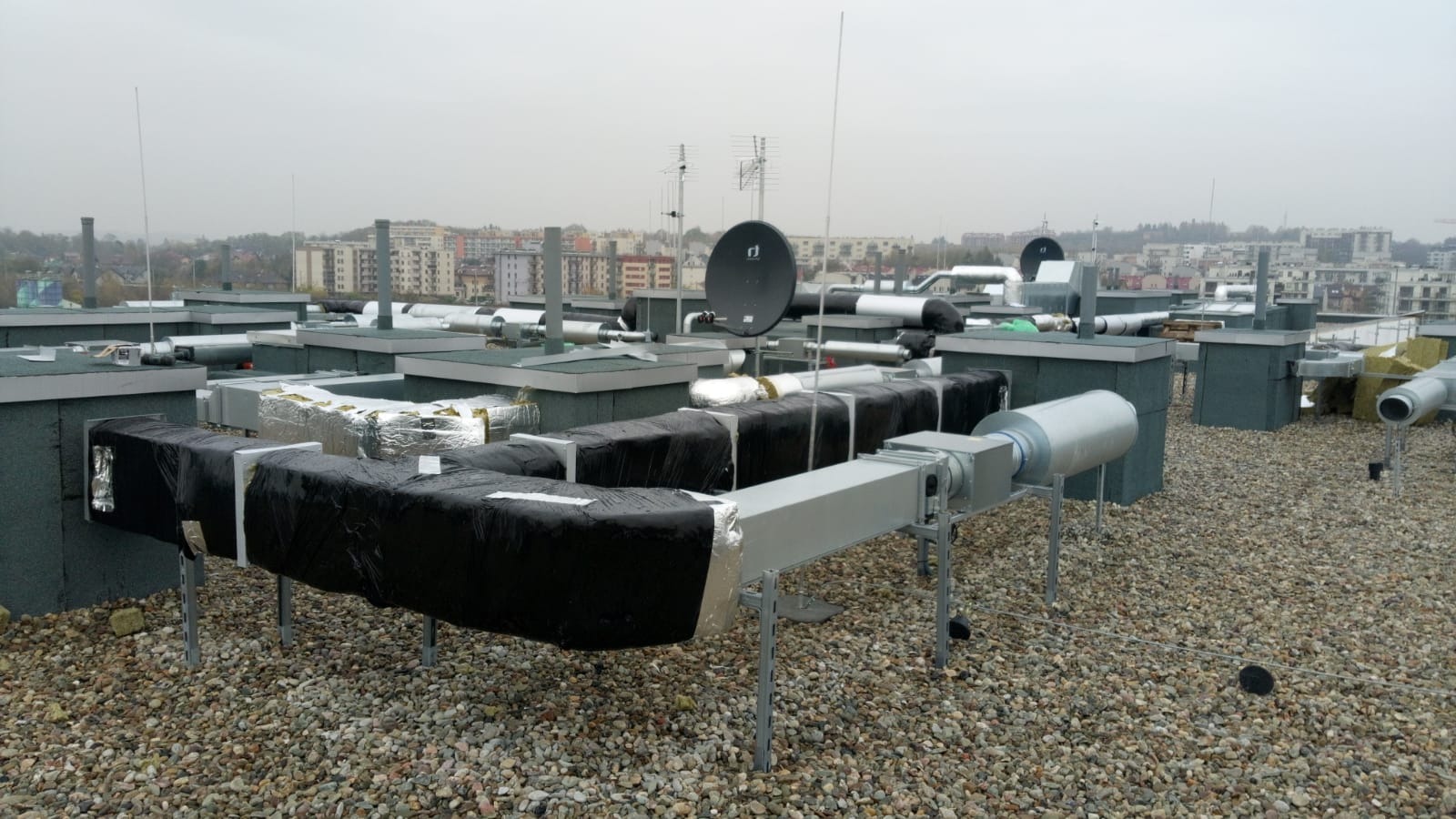 Instalacja wentylacyjna z czarną izolacją na dachu budynku, widoczne przewody wentylacyjne, elementy montażowe i tło miejskiej zabudowy w pochmurny dzień.