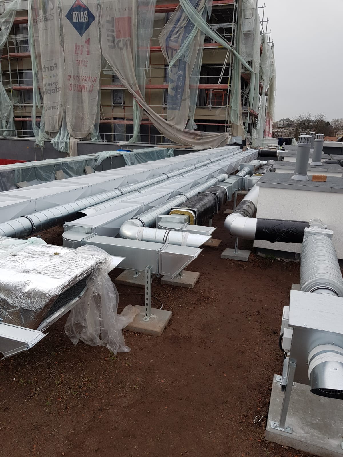 Zewnętrzna instalacja wentylacyjna na dachu budynku w trakcie budowy, widoczne długie, srebrne kanały wentylacyjne, niektóre zaizolowane, oraz rusztowania z plandekami w tle.