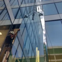 Pracownik w uprzęży, montuje dużą szklaną taflę na wysokościowcu, używając przyssawek i lin.
