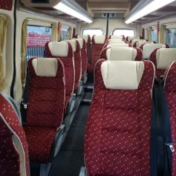 Wnętrze autobusu z rzędami bordowych foteli w białe i pomarańczowe kropki, beżowe zagłówki, czarne pasy bezpieczeństwa i jasne oświetlenie sufitowe.