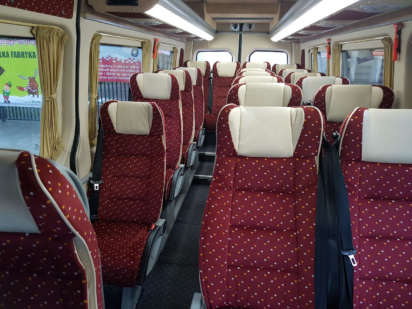 Wnętrze autobusu z rzędami bordowych foteli w białe i pomarańczowe kropki, beżowe zagłówki, czarne pasy bezpieczeństwa i jasne oświetlenie sufitowe.