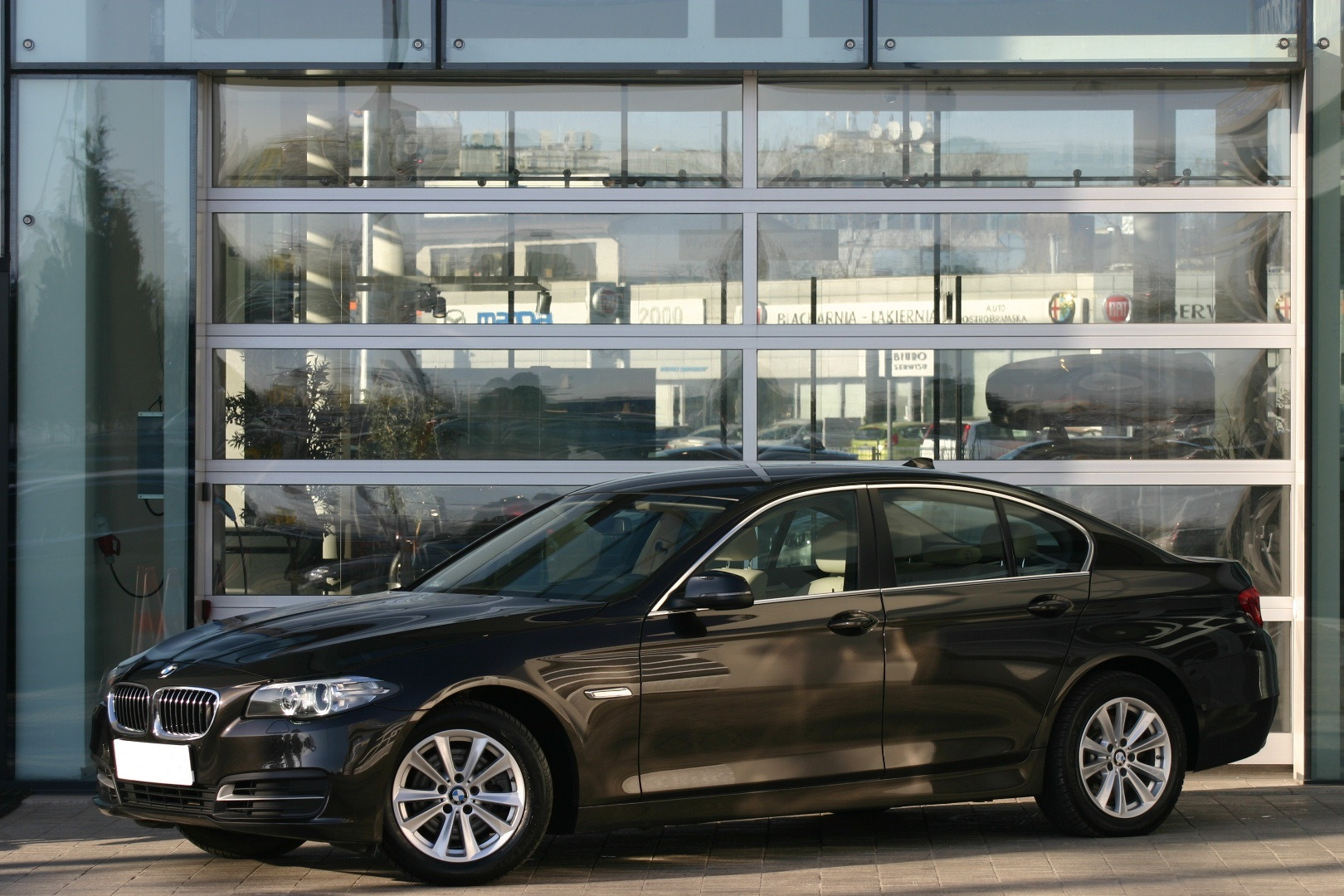 Czarny samochód BMW serii 5 na tle przeszklonej ściany serwisu samochodowego z widocznymi logami różnych marek w tle.