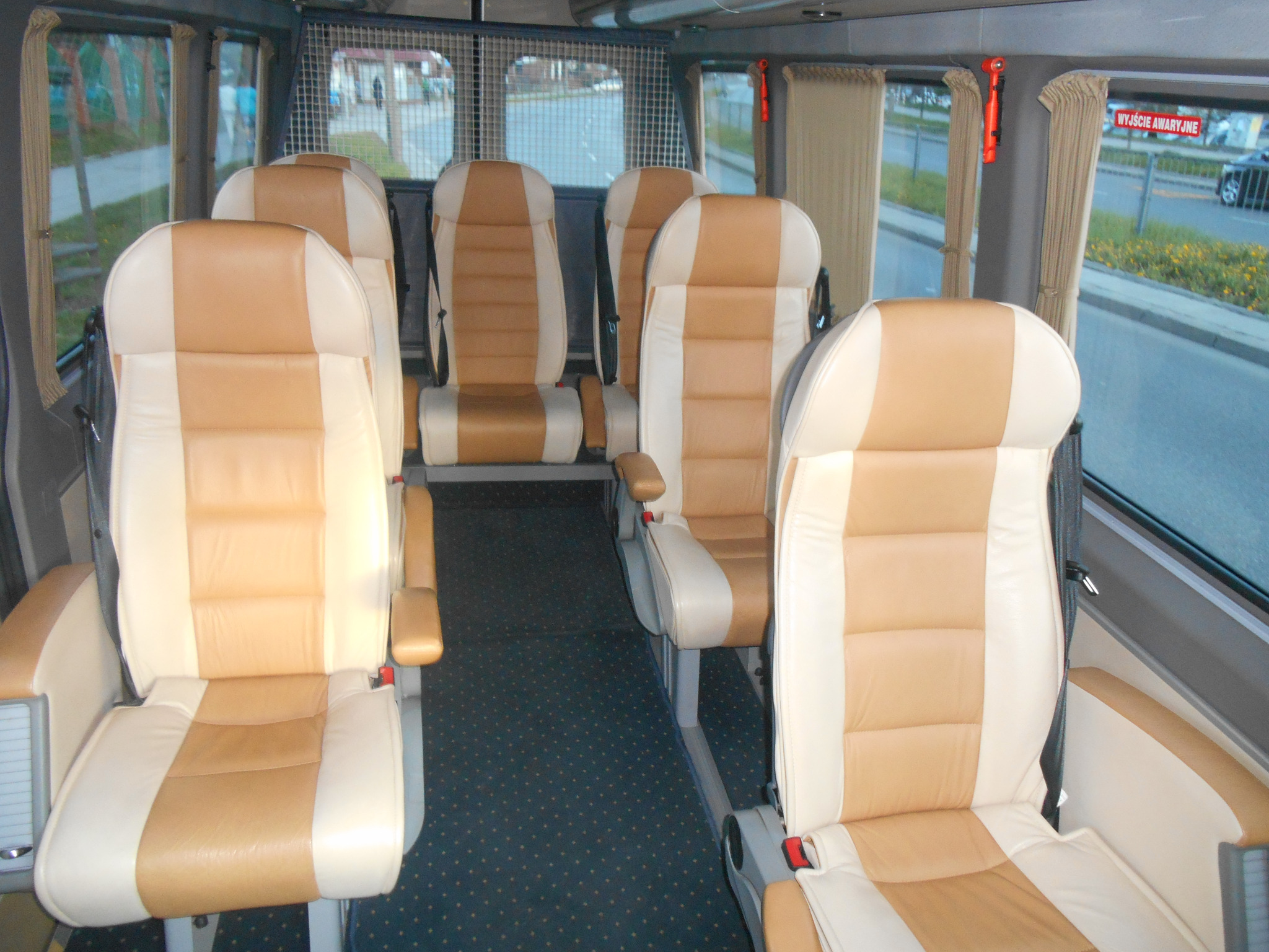 Wnętrze 9-osobowego busa VIP z beżowo-kremowymi skórzanymi fotelami, widok z przodu kabiny, ciemna podłoga w kropki, widoczne zasłony w oknach i czerwony młotek bezpieczeństwa.