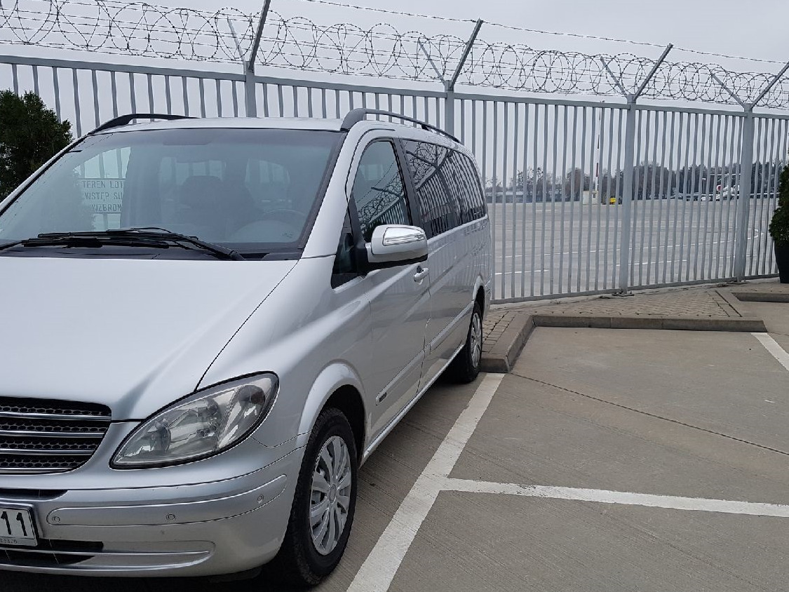 Srebrny van Mercedes zaparkowany na betonowym parkingu z białymi liniami, w tle wysokie metalowe ogrodzenie zwieńczone drutem kolczastym.