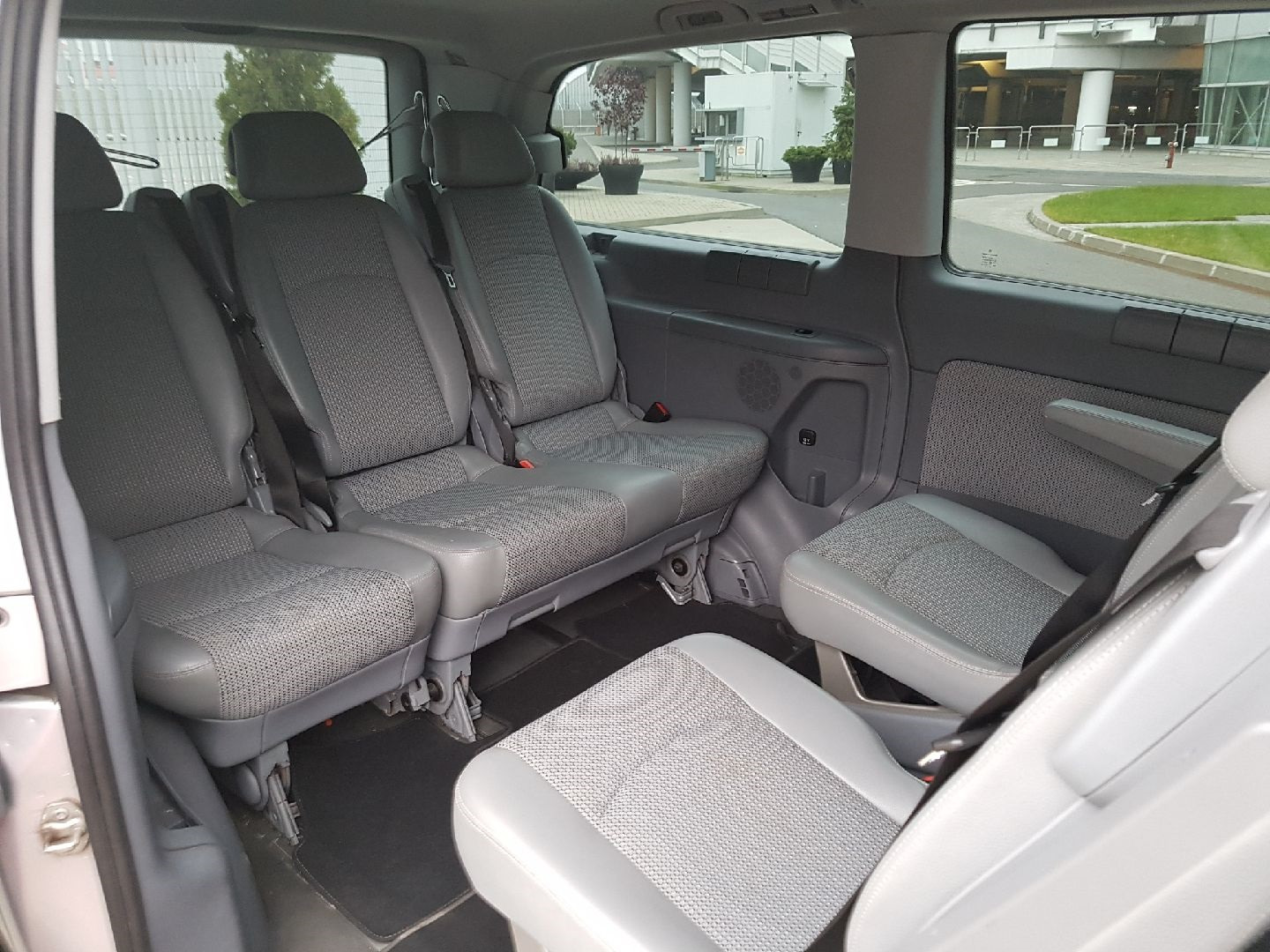 Wnętrze busa z trzema rzędami szarych foteli, widok z boku na puste siedzenia pasażerskie z zagłówkami, czarną wykładziną podłogową i widokiem na budynek z zewnątrz.