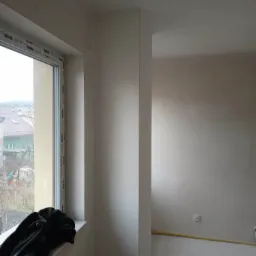 Wnętrze w trakcie prac wykończeniowych: gładkie, jasne ściany, widoczne okno z profilem Rehau, na parapecie czarna kurtka, żółty przewód przy podłodze, gniazdko elektryczne na ścianie.
