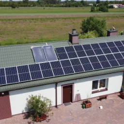 Widok z lotu ptaka na dom z panelami słonecznymi i kolektorami słonecznymi na dachu z zielonej blachy, na tle zielonego pola i lasu.