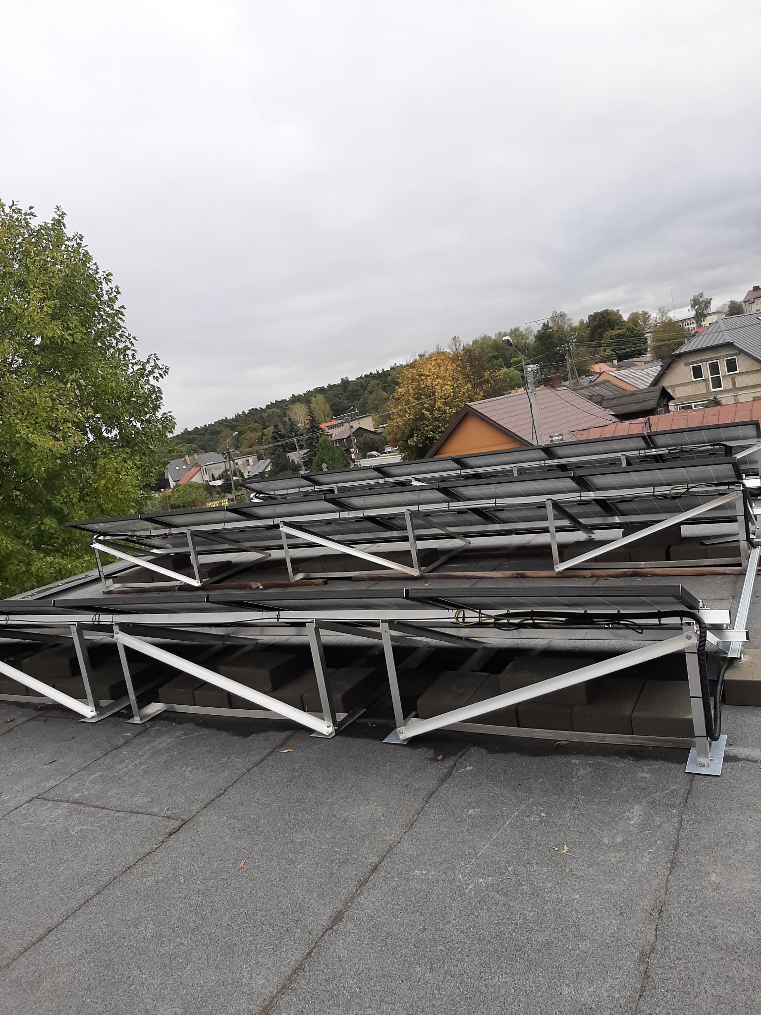 Rząd paneli słonecznych na płaskim dachu, z widoczną aluminiową konstrukcją wsporczą i elementami mocującymi, w tle zabudowa miejska pod pochmurnym niebem.