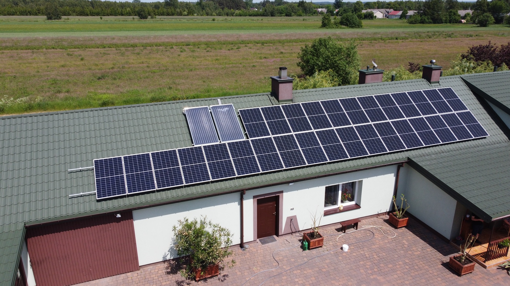 Widok z lotu ptaka na dom z panelami słonecznymi i kolektorami słonecznymi na dachu z zielonej blachy, na tle zielonego pola i lasu.