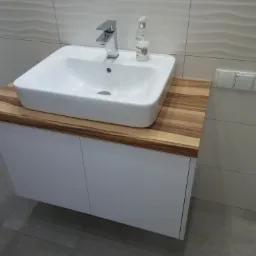 Biała umywalka nablatowa z chromowaną baterią i dozownikiem mydła, osadzona na blacie z drewna egzotycznego, zamontowana na białej szafce łazienkowej.