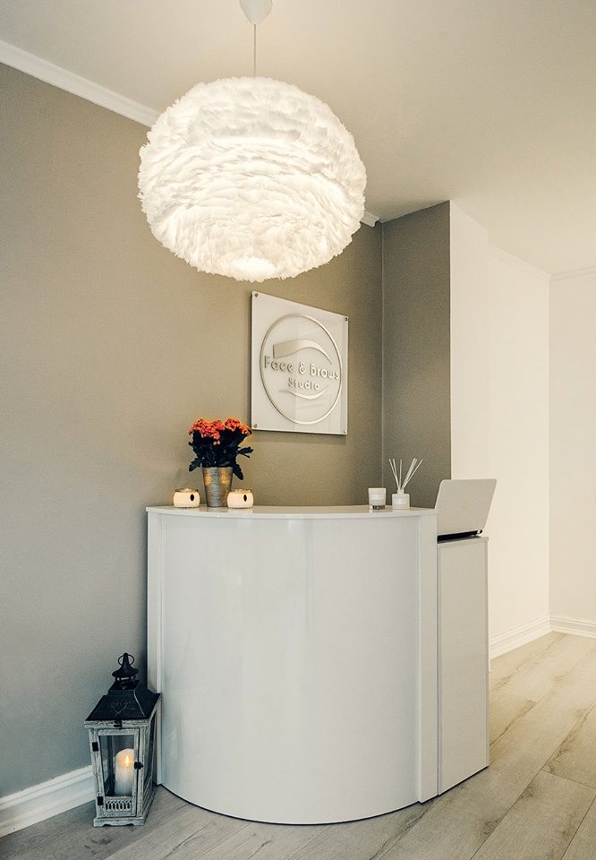 Biała lada recepcyjna z zaokrąglonym frontem w salonie kosmetycznym, ozdobiona kwiatami w metalowym wiaderku, świecami i lampionem podłogowym, nad nią wisi puszysta lampa i logo salonu.