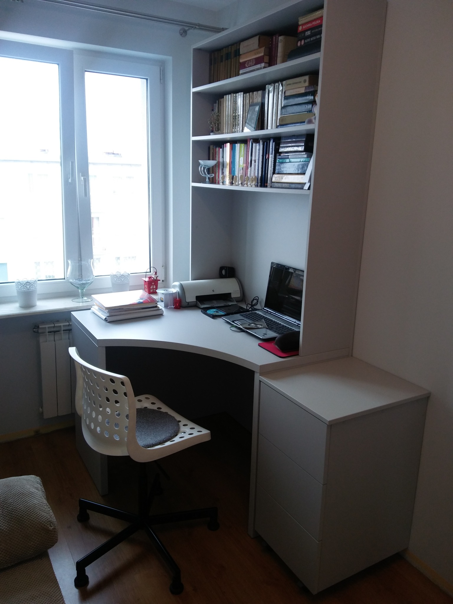 Ergonomiczne biurko narożne z szufladami i regałem na książki, z laptopem i drukarką, ustawione przy oknie.