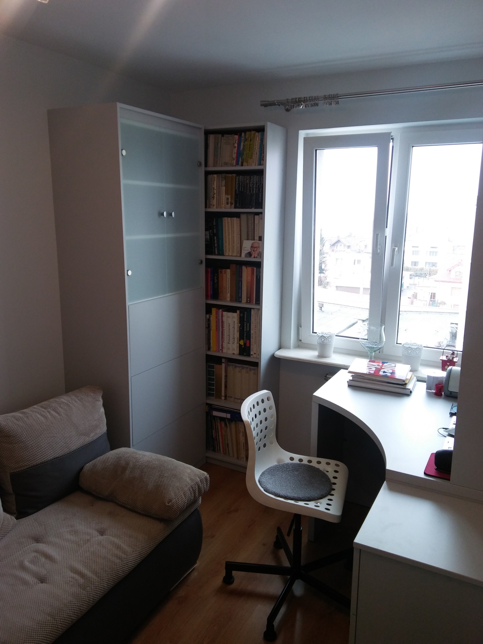 Pokój z białymi meblami: szafa z częściowo przeszklonymi drzwiami, regał z książkami, biurko przy oknie i sofa z szarą tapicerką.