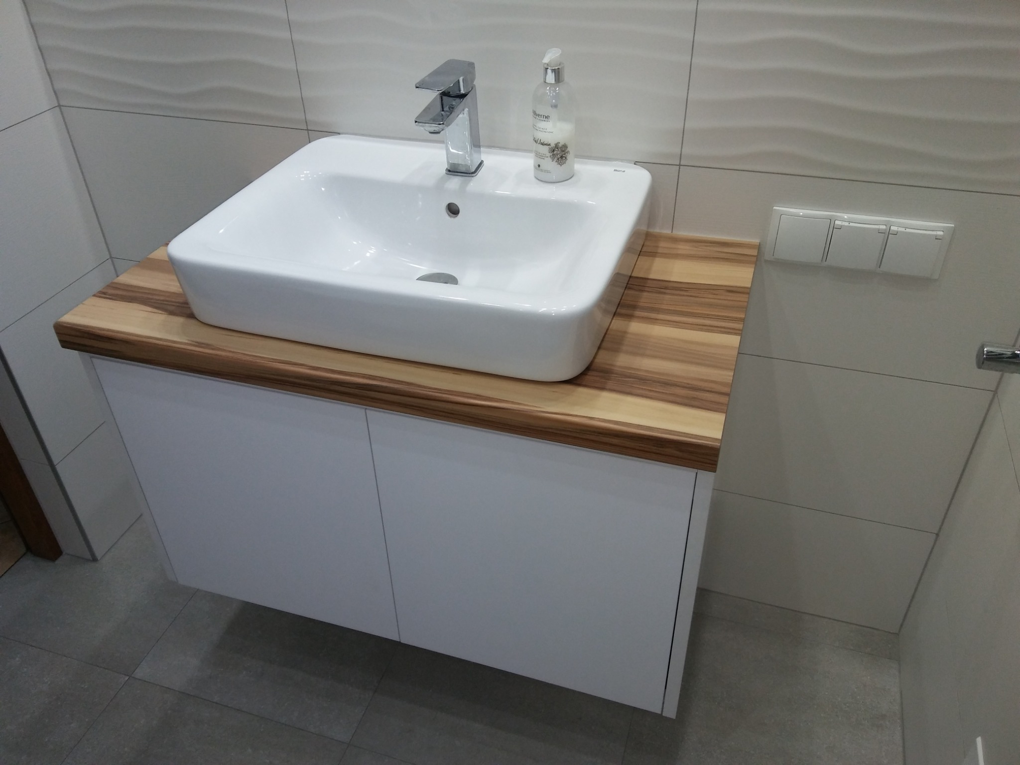 Biała umywalka nablatowa z chromowaną baterią i dozownikiem mydła, osadzona na blacie z drewna egzotycznego, zamontowana na białej szafce łazienkowej.